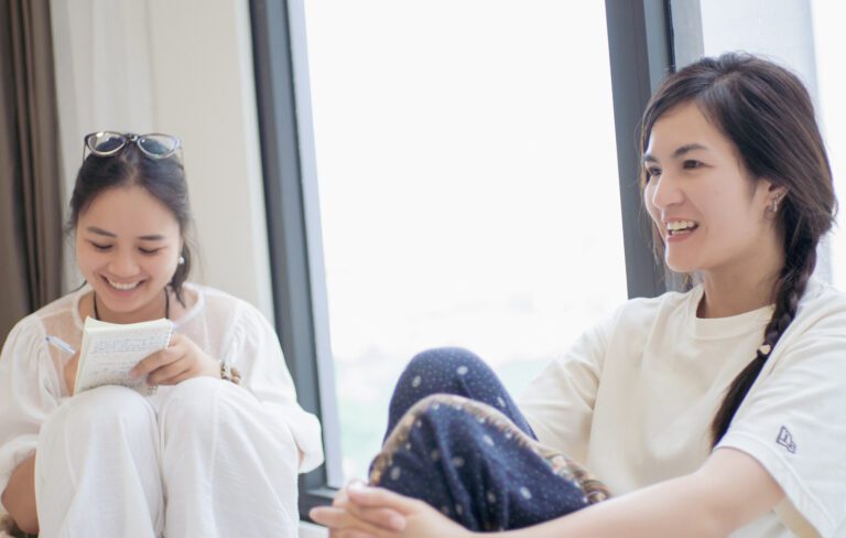 Khóa Học Gieo Duyên Cùng Chuông Xoay Himalaya 17-18/08/2024 27 Hai cô gái trò chuyện vui vẻ cạnh cửa sổ.